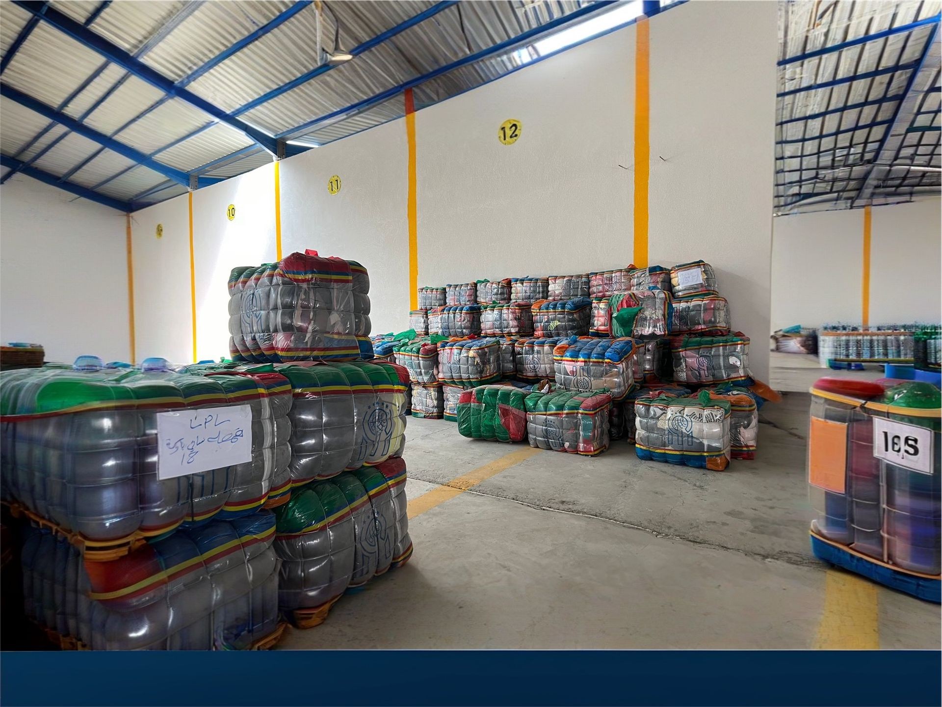 Warehouse interior with stacks of colorfully wrapped palletized goods against white walls.